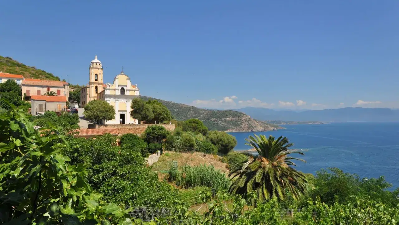 Vue d'un village corse avec une église jaune, des maisons aux toits rouges, des arbres verdoyants et la mer bleue. Un paysage typique de mare e monti corse.
