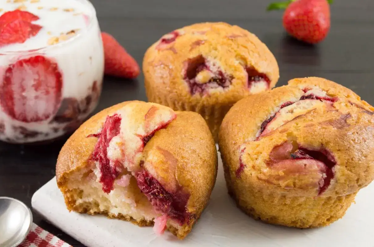 Pyszne muffinki z truskawkami, jeden nadgryziony, obok deser z jogurtem i owocami.