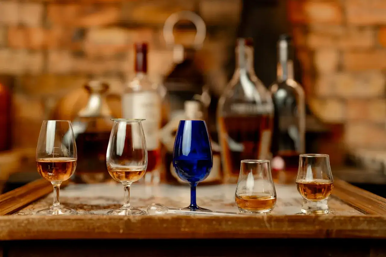 Verkostung von **blended malt whisky**: F&uuml;nf Gl&auml;ser mit bernsteinfarbener Fl&uuml;ssigkeit stehen auf einem Holztisch vor Flaschen und einer Ziegelwand.