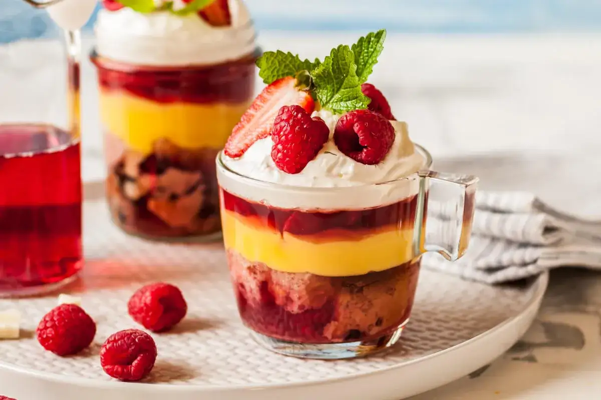 Deliziosa zuppa inglese ricetta in coppetta, con strati di pan di Spagna, crema pasticcera, frutti di bosco e panna montata, guarnita con fragole e lamponi.