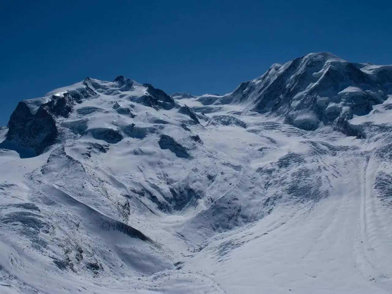 Vastes &eacute;tendues enneig&eacute;es sous un ciel bleu intense, &eacute;voquant l'ascension du Mont Rose. Des glaciers sculptent le paysage montagneux.