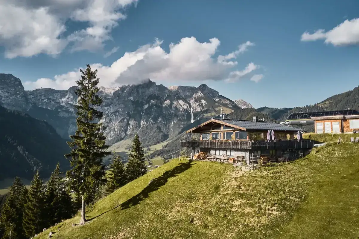 Ein Holzhaus mit Terrasse und Bergblick in Aja, Werfenweng. Im Hintergrund majestätische Gipfel und eine Seilbahnstation.