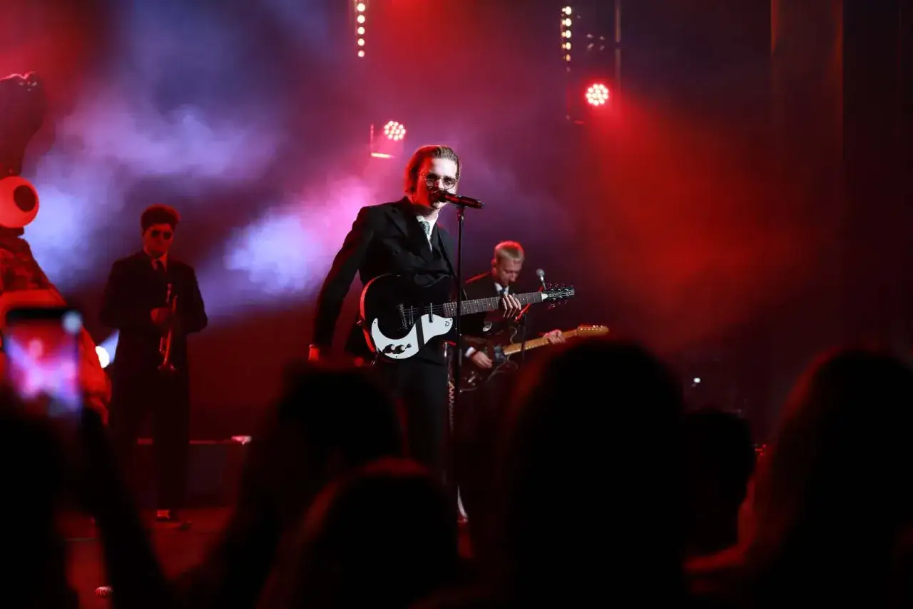 Bracia Kacperczyk na scenie, wokalista z gitarą, w tle trębacz i gitarzysta. Czerwone światła i dym tworzą atmosferę koncertu.