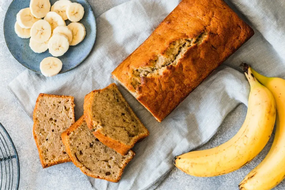 Pyszne ciasto z bananem, pokrojone na kromki, obok świeże banany i talerz z plasterkami.