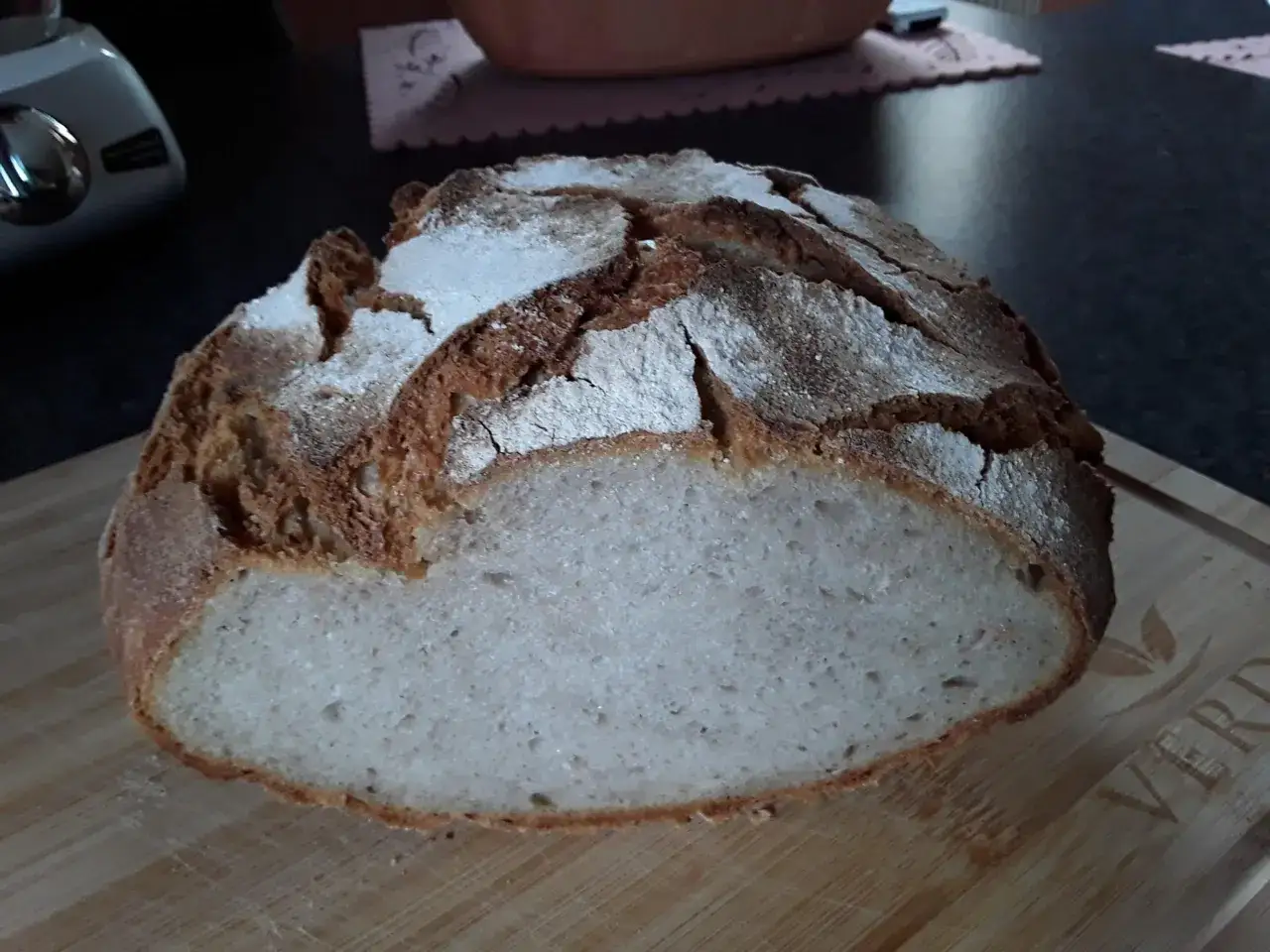 Ein frisch gebackenes Buchweizenbrot mit Sauerteig liegt auf einem Holzbrett. Die Kruste ist knusprig und mit Mehl best&auml;ubt.