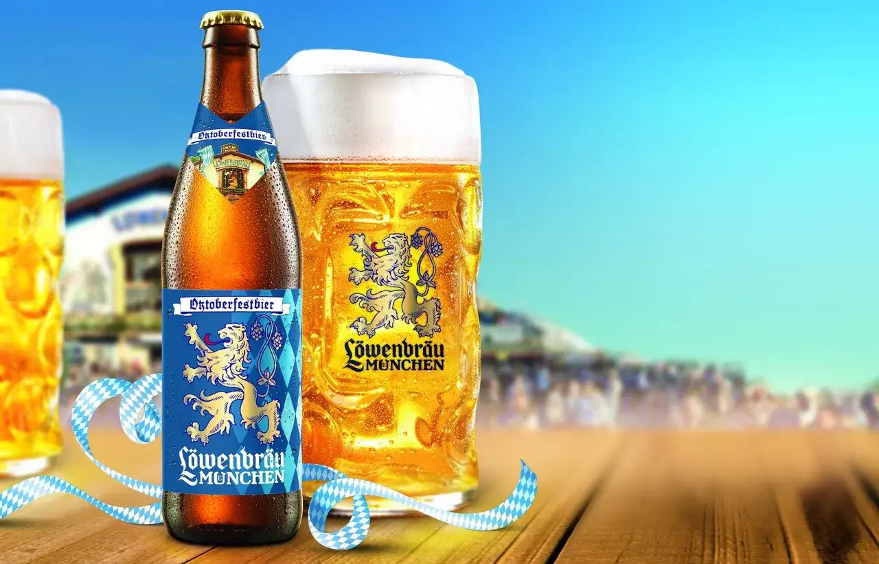 Birre dell'Oktoberfest L&ouml;wenbr&auml;u M&uuml;nchen, con un bicchiere pieno e una bottiglia su un tavolo di legno, sullo sfondo di una folla festante.