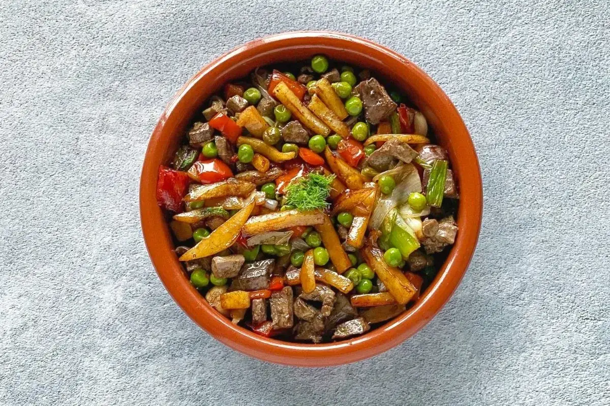 Un sabroso frito mallorqu&iacute;n con trozos de carne, patatas fritas, guisantes y pimientos rojos en una cazuela de barro.