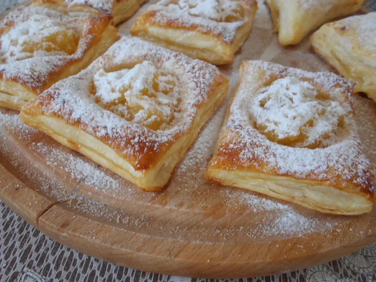 Słodkie ciasto francuskie z jabłkami, posypane cukrem pudrem, prezentuje się apetycznie na drewnianej desce.