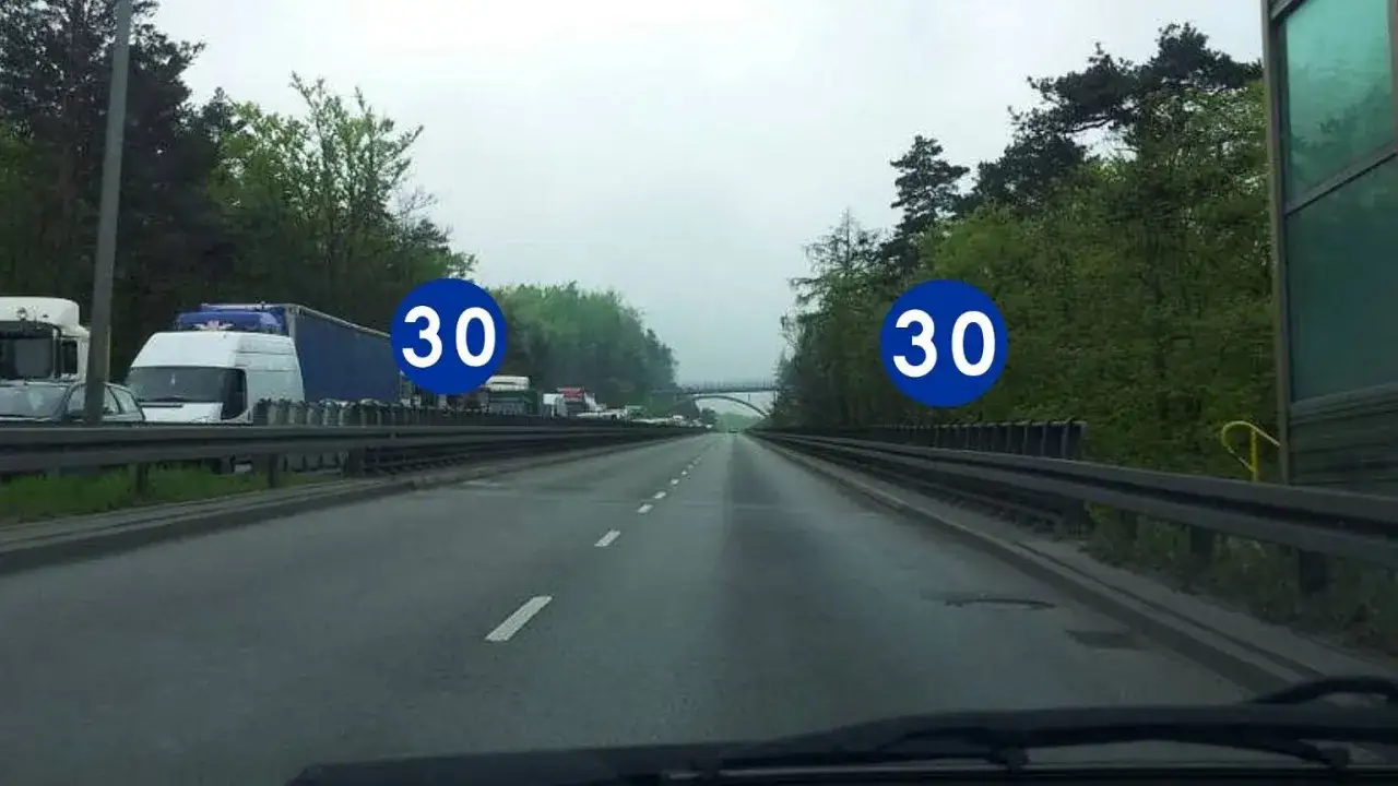 Na autostradzie znaki ograniczają prędkość do 30 km/h. Widać sznur ciężarówek i samochodów.