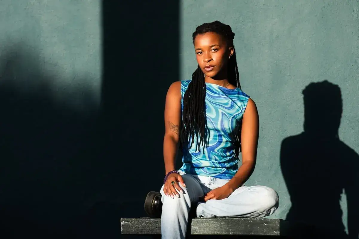 A woman with dreadlocks sits cross-legged in the broad lighting of a sunny day, her blue patterned top and light jeans illuminated against a dark wall.