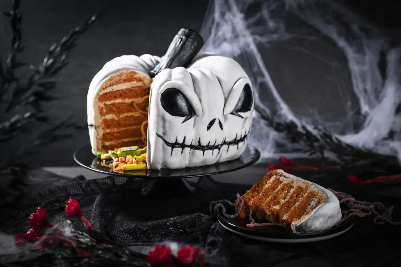 Gruselige Halloween-Torte in K&uuml;rbisform mit grimmigem Gesicht. Ein St&uuml;ck des Kuchens mit orangefarbenen Schichten liegt daneben.