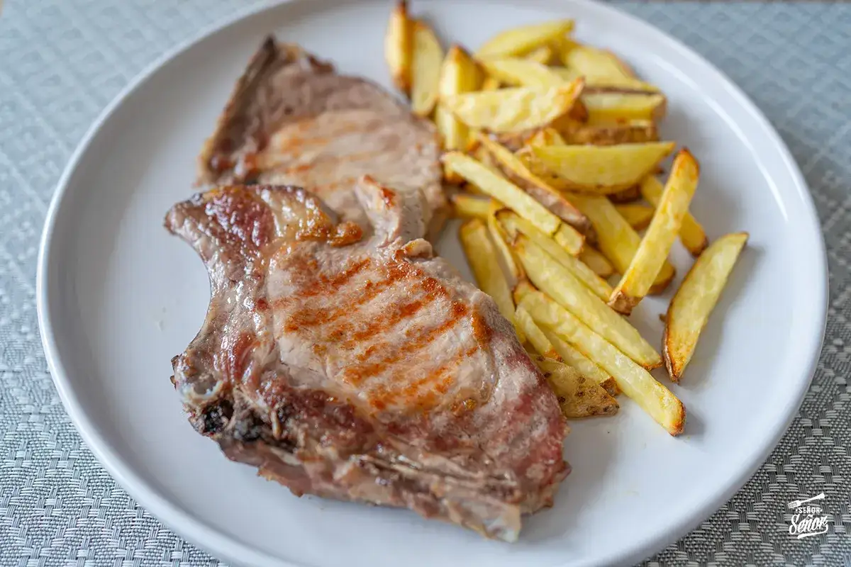 Dos jugosas chuletas de cerdo a la parrilla acompa&ntilde;adas de patatas fritas crujientes en un plato blanco.