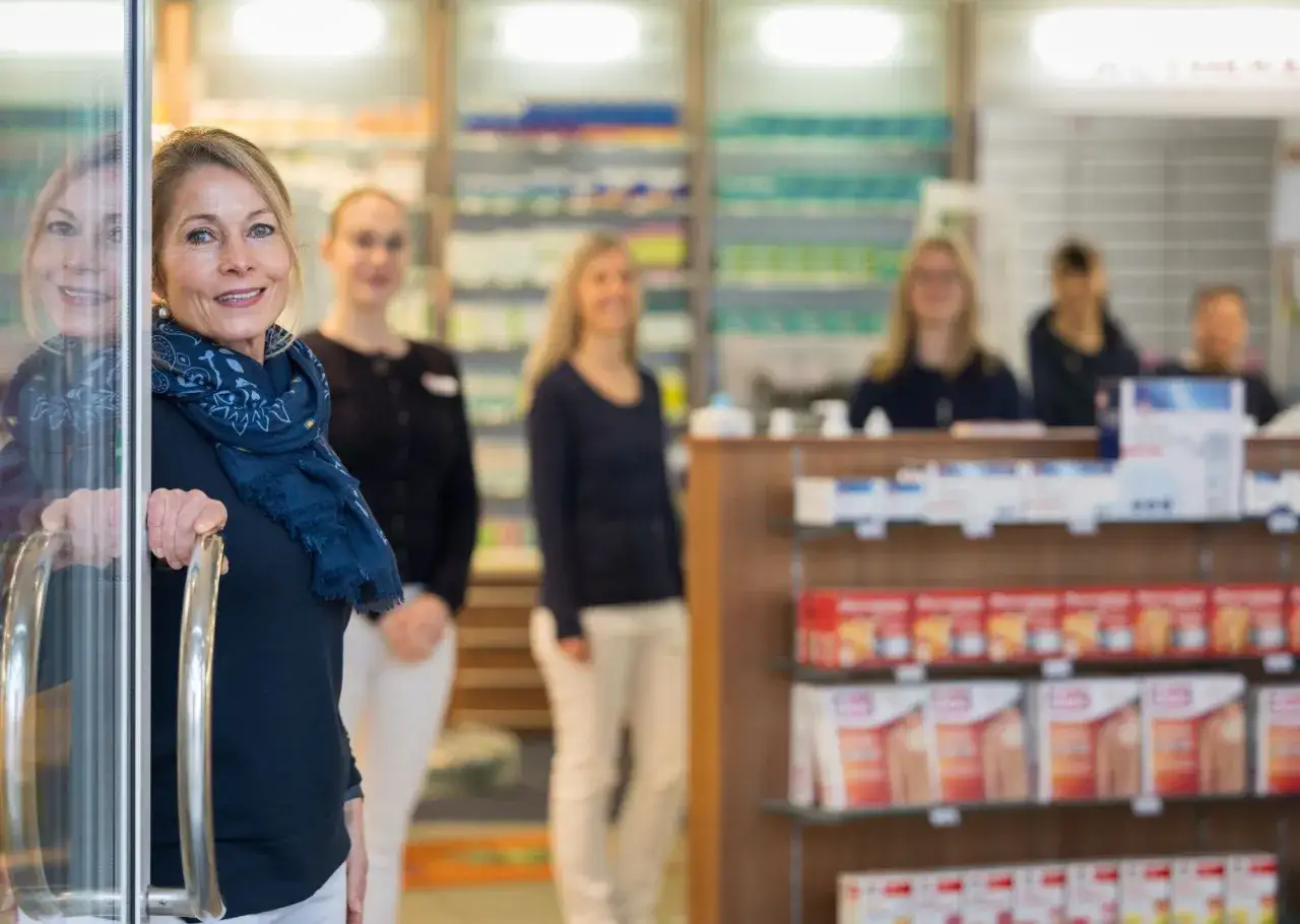 Frau betritt Apotheke, fragt nach ellaOne Nebenwirkungen. Mehrere Apothekerinnen im Hintergrund.