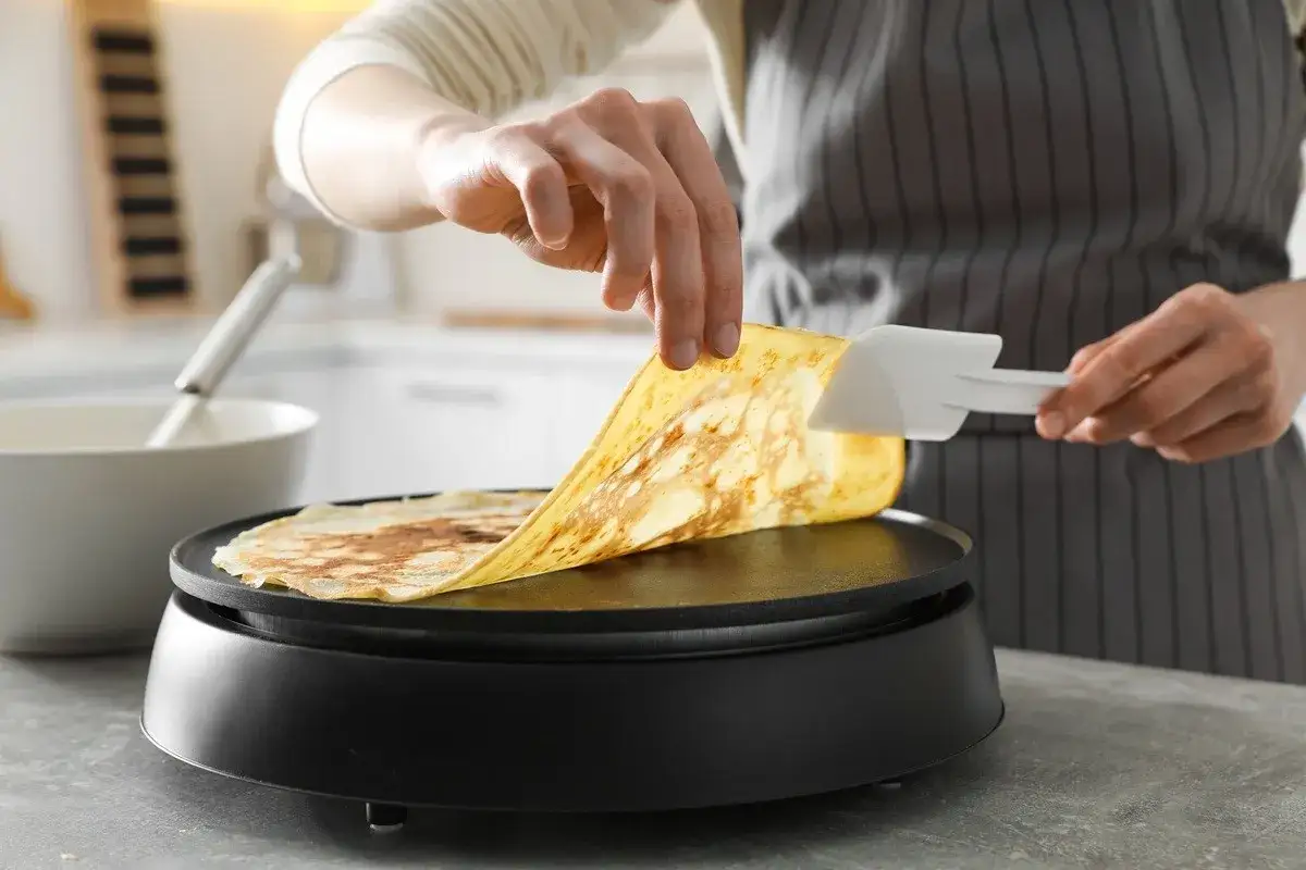 Une main soulève une crêpe dorée d'une crêpière Tefal, prête pour le nettoyage.