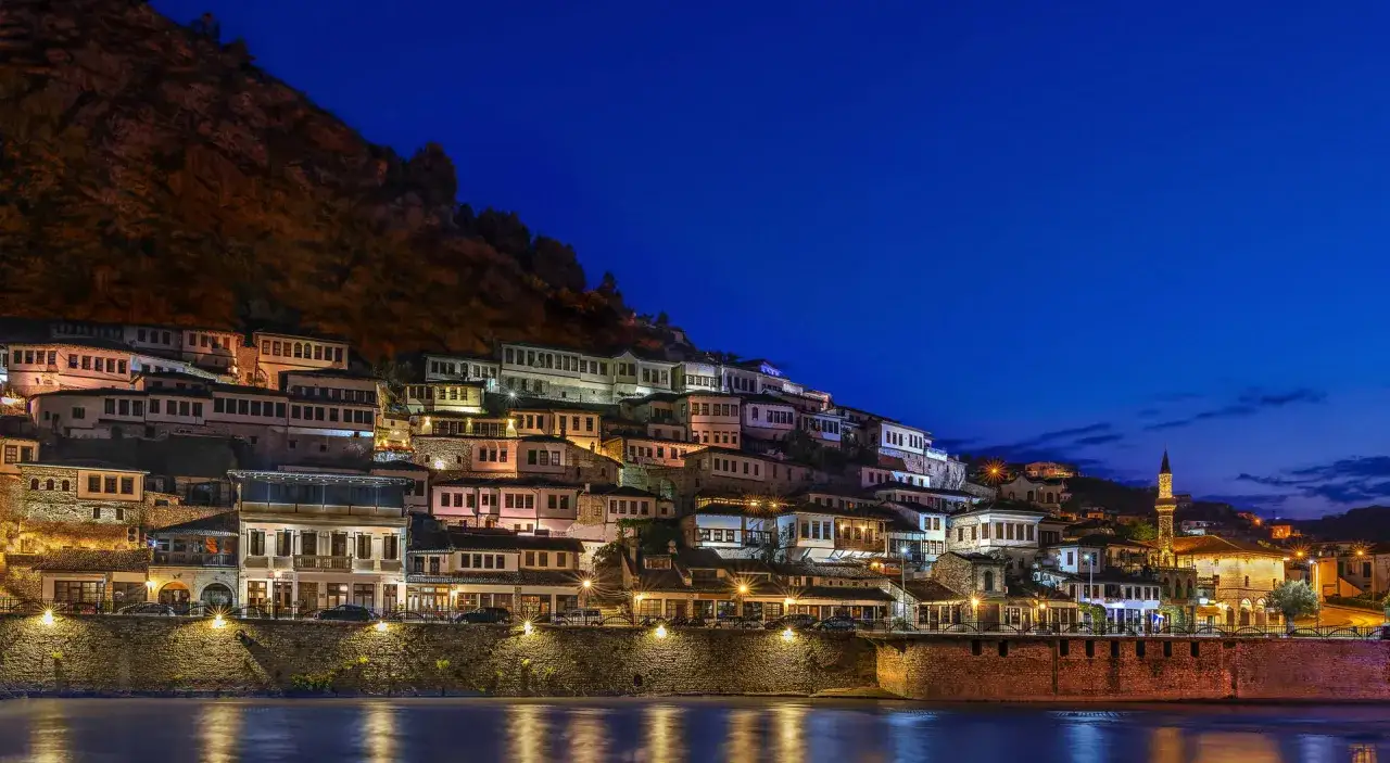 Albania, styczeń. Nocą oświetlone domy na zboczu g&oacute;ry w Berat, odbijające się w rzece.