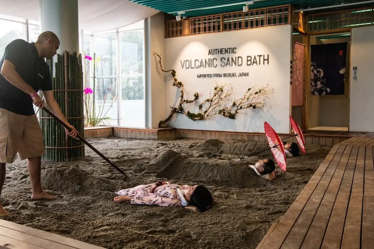 A man rakes sand for a volcanic sand bath. Two people lie buried in the sand, with red umbrellas over their heads.