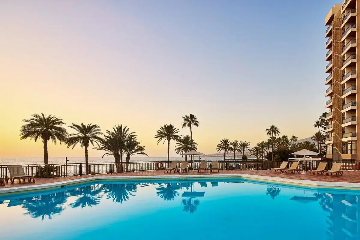 Basen z turkusową wodą i odbijającymi się palmami, w tle hotel i ocean. Idealne miejsce na wakacje na Playa de las Américas.