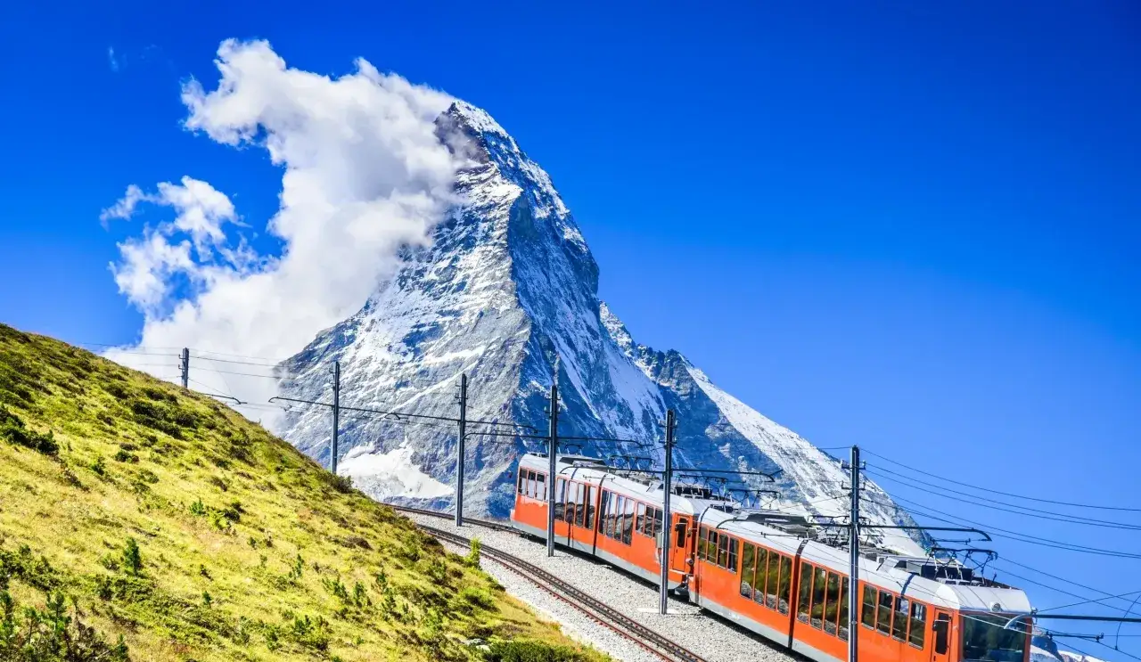 Pomarańczowy pociąg Glacier Express pnie się po zielonym zboczu w kierunku ośnieżonego szczytu Matterhorn.