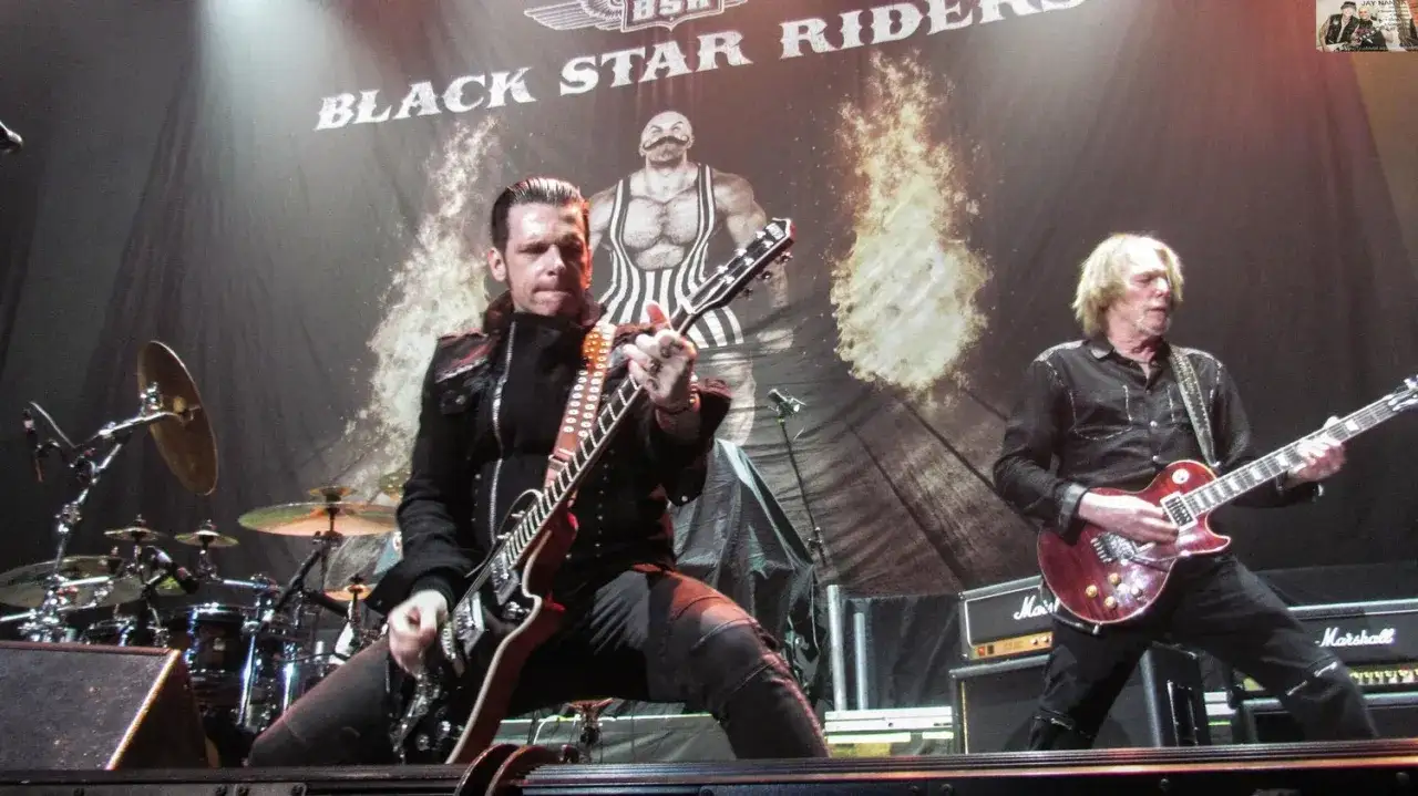 Les **Black Star Riders** en concert, deux guitaristes sur scène devant une bannière enflammée et un homme musclé.
