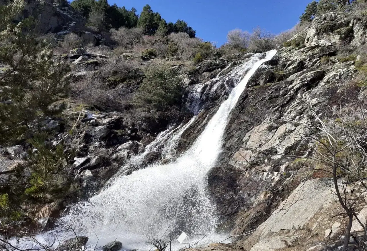 Cascada en San Mamés, Madrid. El agua cae con fuerza por las rocas, rodeada de vegetación y un cielo azul intenso.