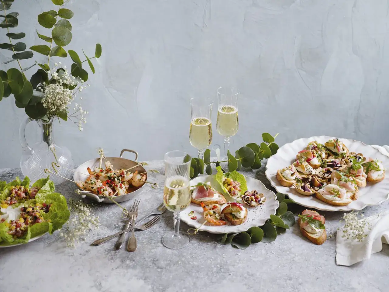 Festliga tilltugg till bubbel: räkor på spett, bruschetta med olika pålägg och sallad i salladsblad. Två glas bubbel står redo.