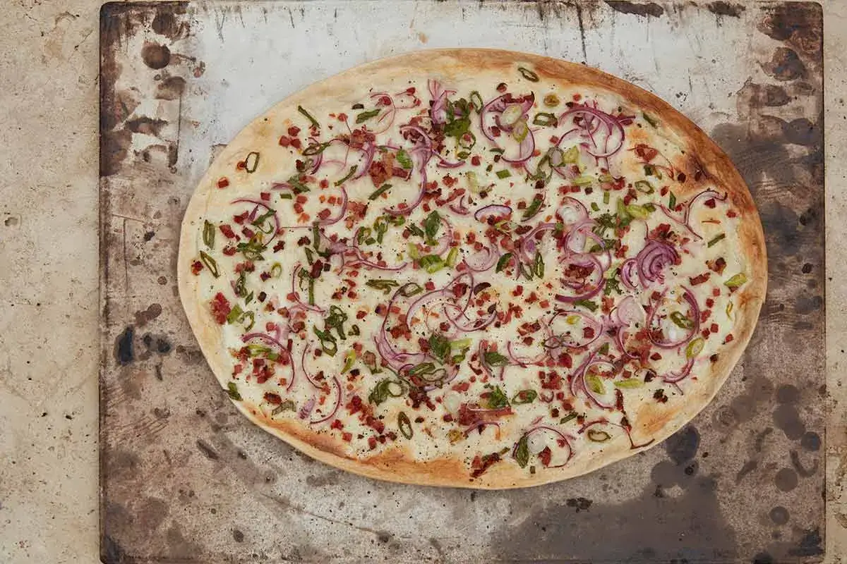 Flammkuchen mit dünnem Teig, belegt mit Crème fraîche, Speck, roten Zwiebeln und Frühlingszwiebeln.