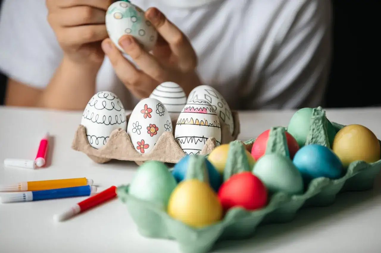 Bambino decora uova con pennarelli, creando disegni colorati. Sul tavolo, uova già dipinte e pennarelli pronti per i prossimi colori per uova di Pasqua.