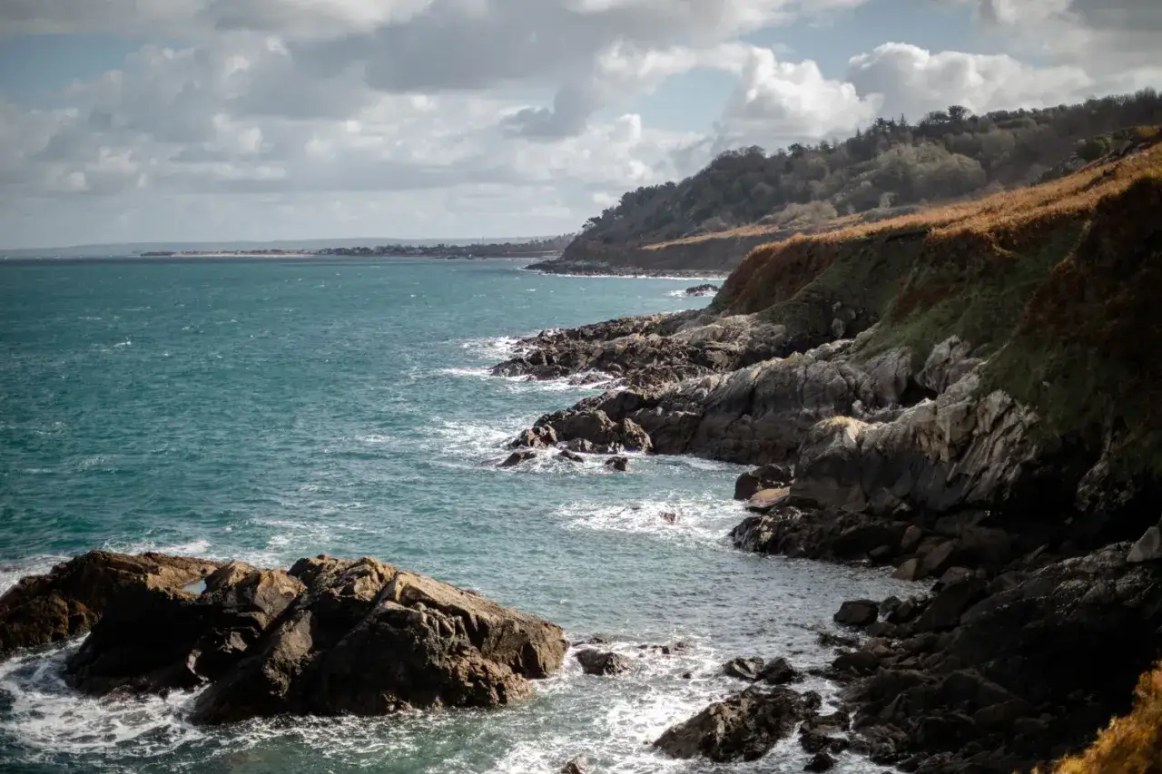 C&ocirc;te rocheuse battue par les vagues de la Manche, avec une colline verdoyante et des nuages dans le ciel.