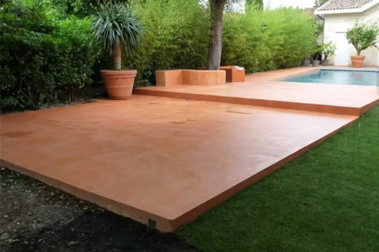 Une belle terrasse en béton coloré ocre, parfaite pour un devis terrasse béton. Entourée de verdure et d'une piscine.