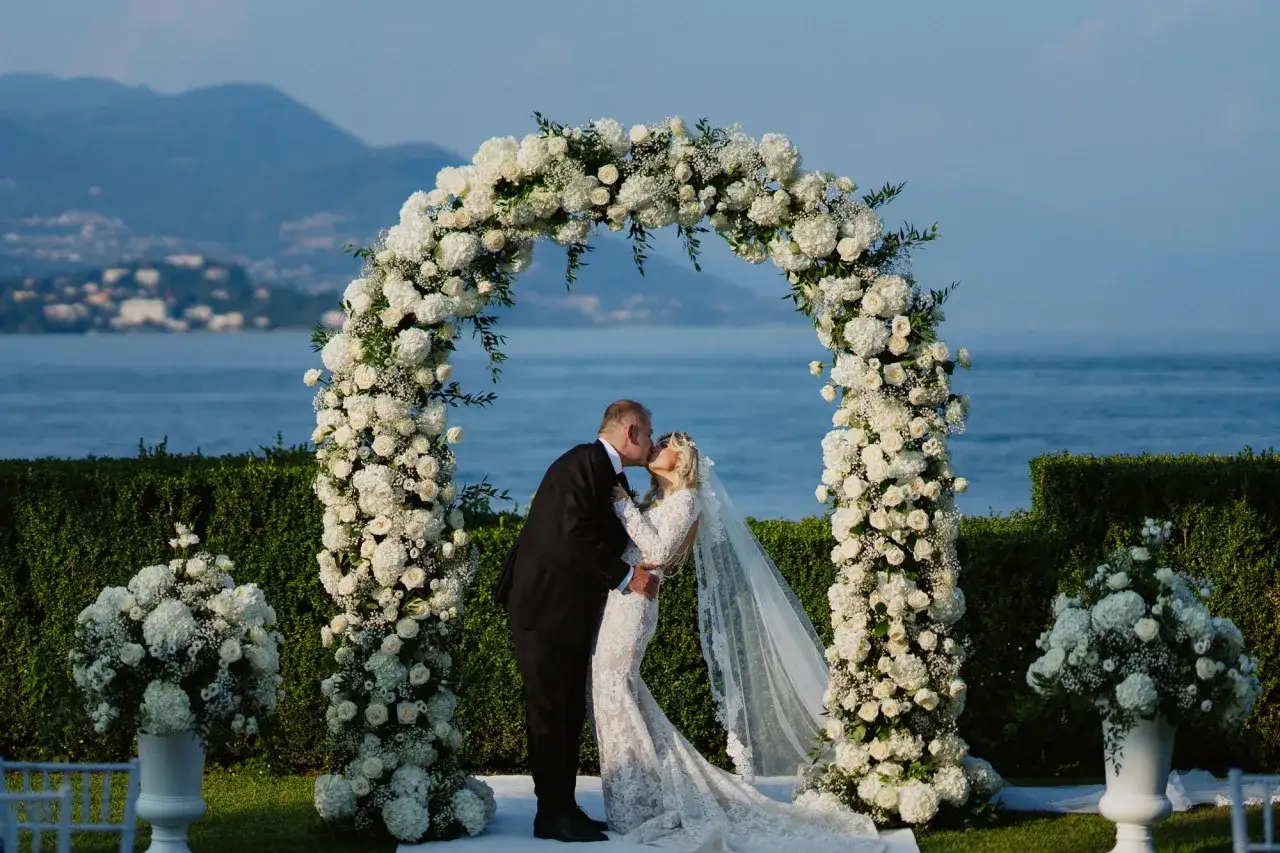 Sposi si baciano sotto un arco fiorito, con il lago e le montagne sullo sfondo. Un luogo da sogno dove festeggiare anniversario matrimonio.