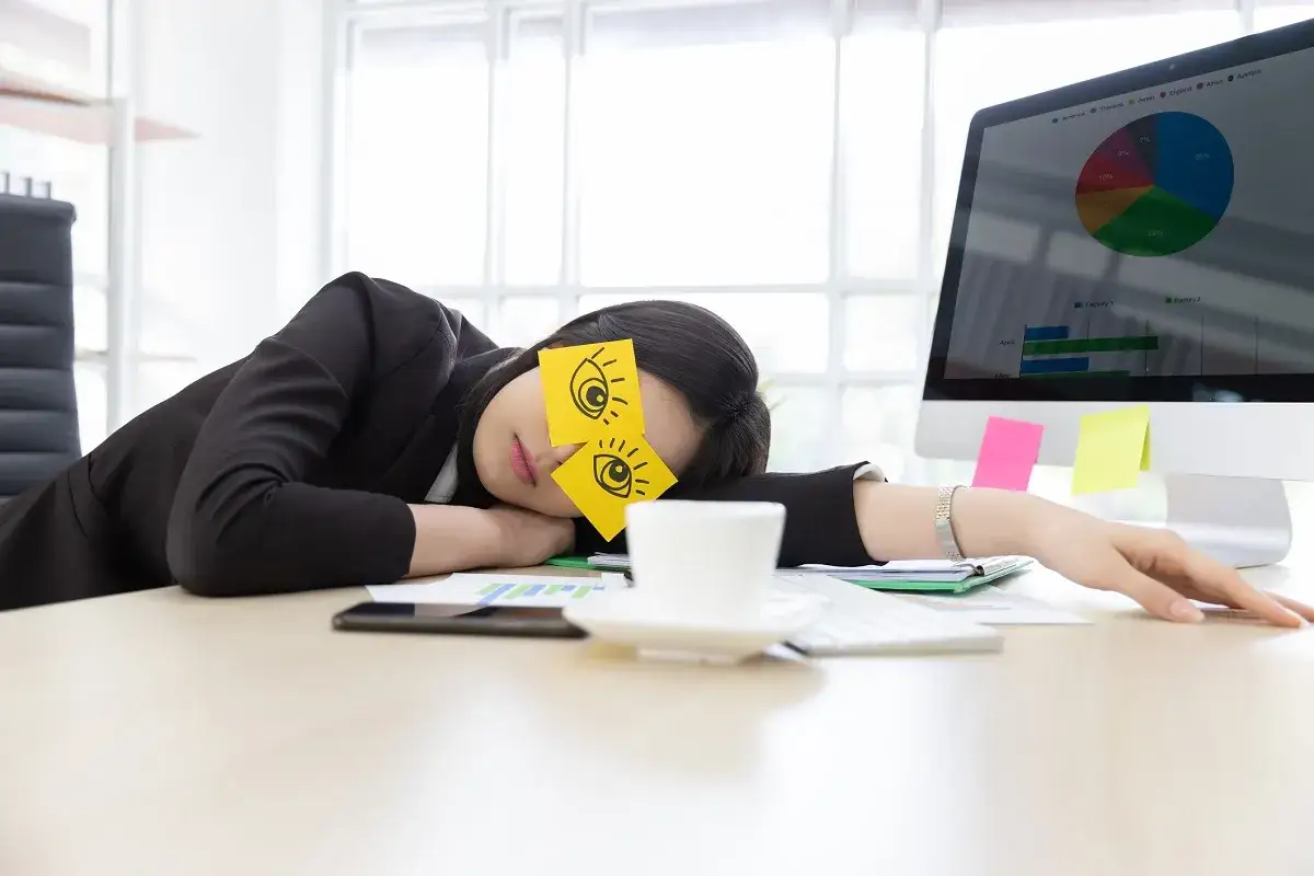 En kvinna sover vid sitt skrivbord med post-it-lappar med &ouml;gon p&aring; &ouml;gonlocken. Det &auml;r verkligen tr&aring;kigt p&aring; jobbet.