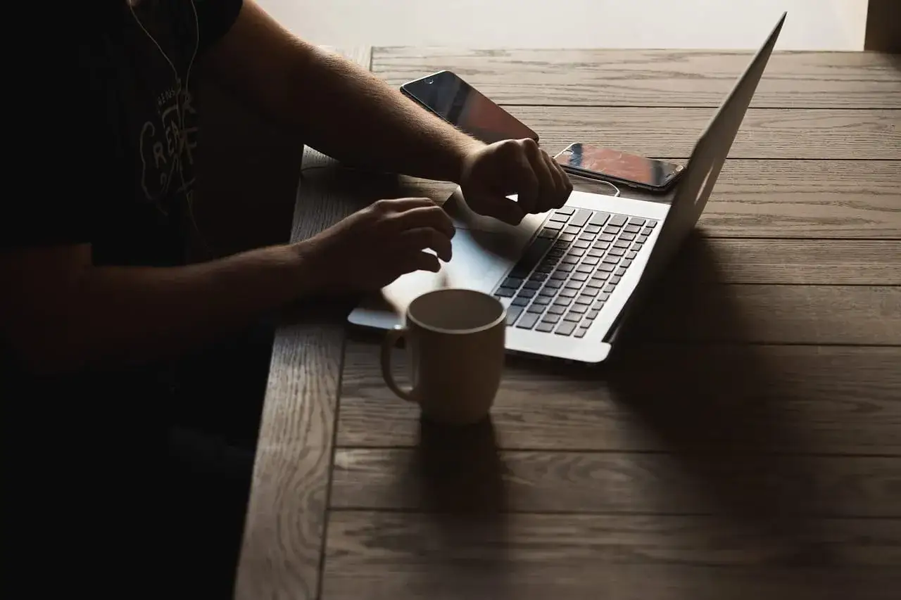 Osoba pracuje na laptopie, obok leżą telefony i kubek. Czy Airbnb jest bezpieczne? Sprawdzanie opinii i warunków przed rezerwacją.