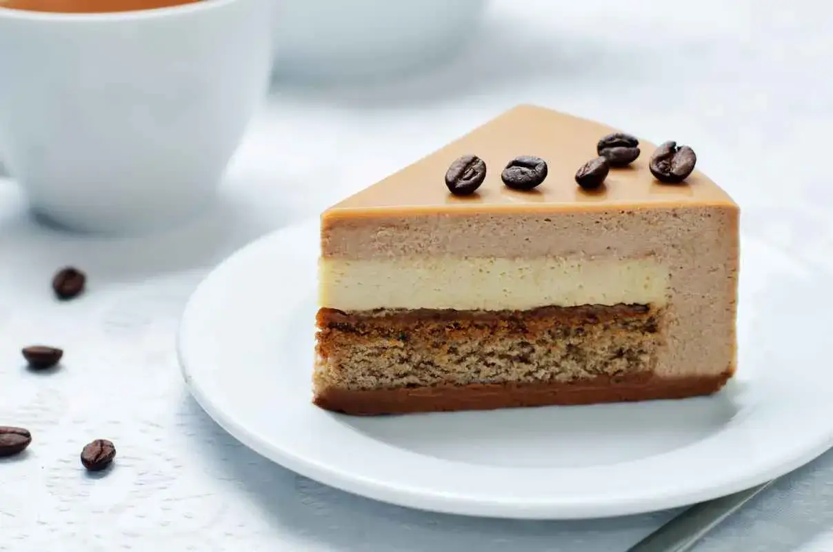 Una fetta di cheesecake al caff&egrave; a pi&ugrave; strati, guarnita con chicchi di caff&egrave;, su un piatto bianco.