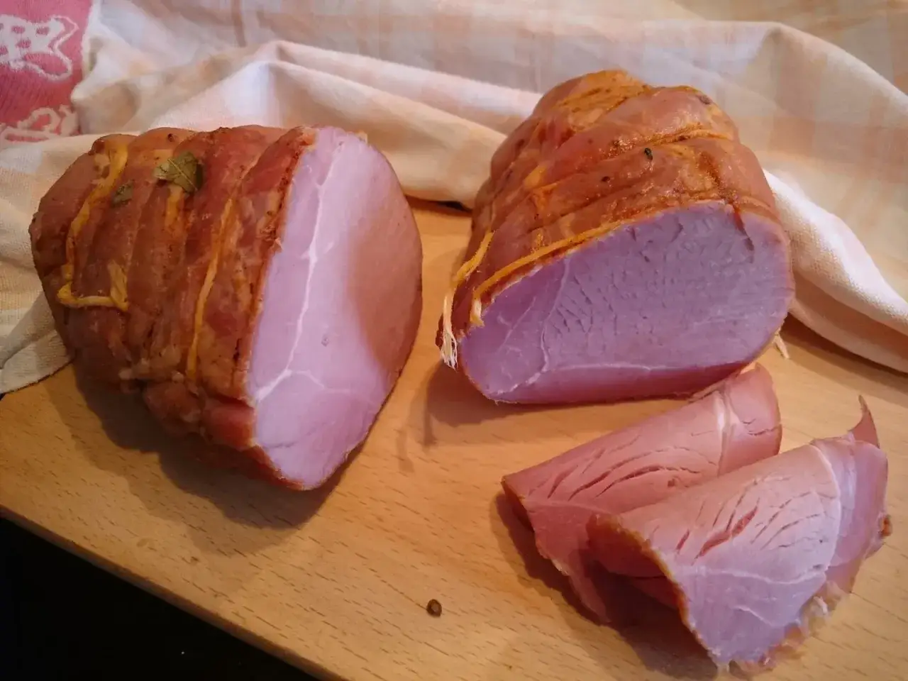 Dwie wędzone szynki, jedna przekrojona, pokazują, jak peklować szynkę do pieczenia. Na desce leżą też plastry mięsa.