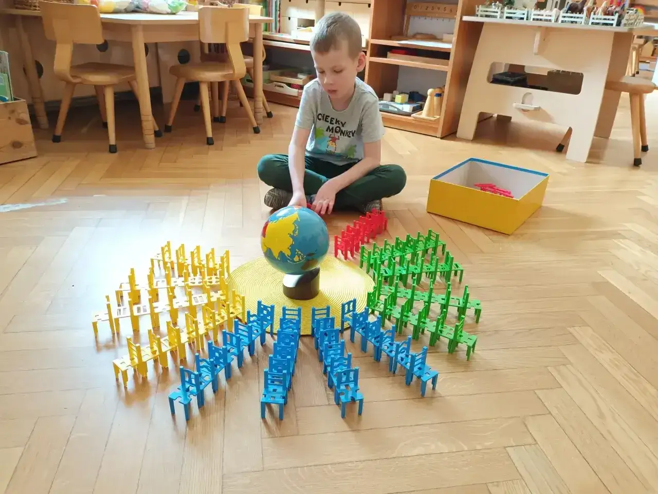 Chłopiec bawi się w przedszkolu Montessori, układając kolorowe krzesełka wokół globusa. To pokazuje, montessori co to za metoda nauczania.