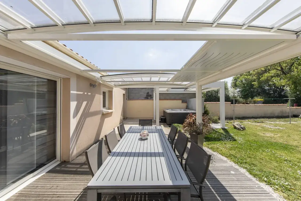 Tavolo da pranzo e sedie sotto una moderna veranda con tetto trasparente, un angolo relax con vasca idromassaggio. Una veranda senza permessi che amplia lo spazio abitativo.