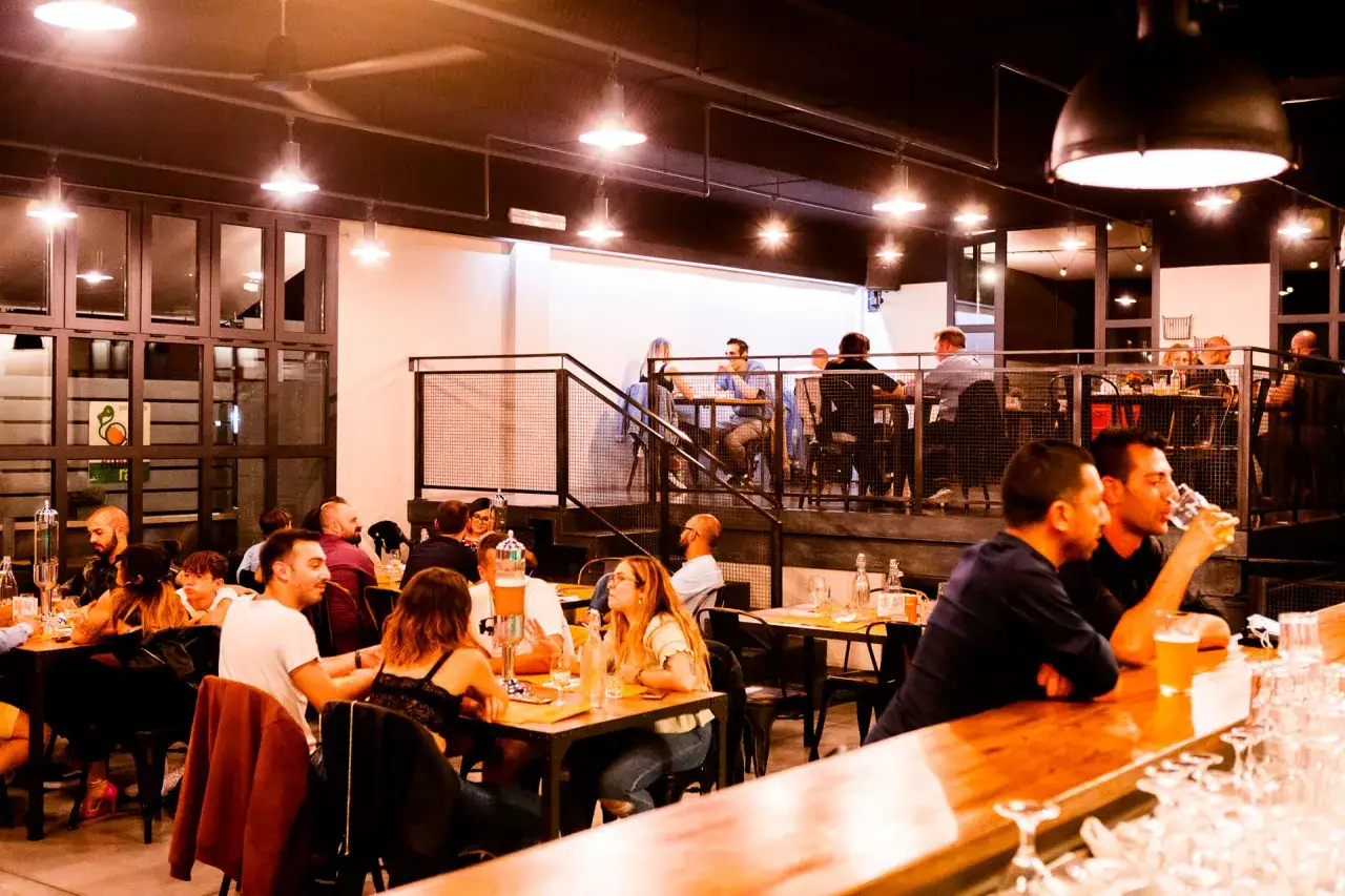 Persone sedute ai tavoli e al bancone del birrificio Rubiu, gustando birra e cibo in un'atmosfera conviviale.