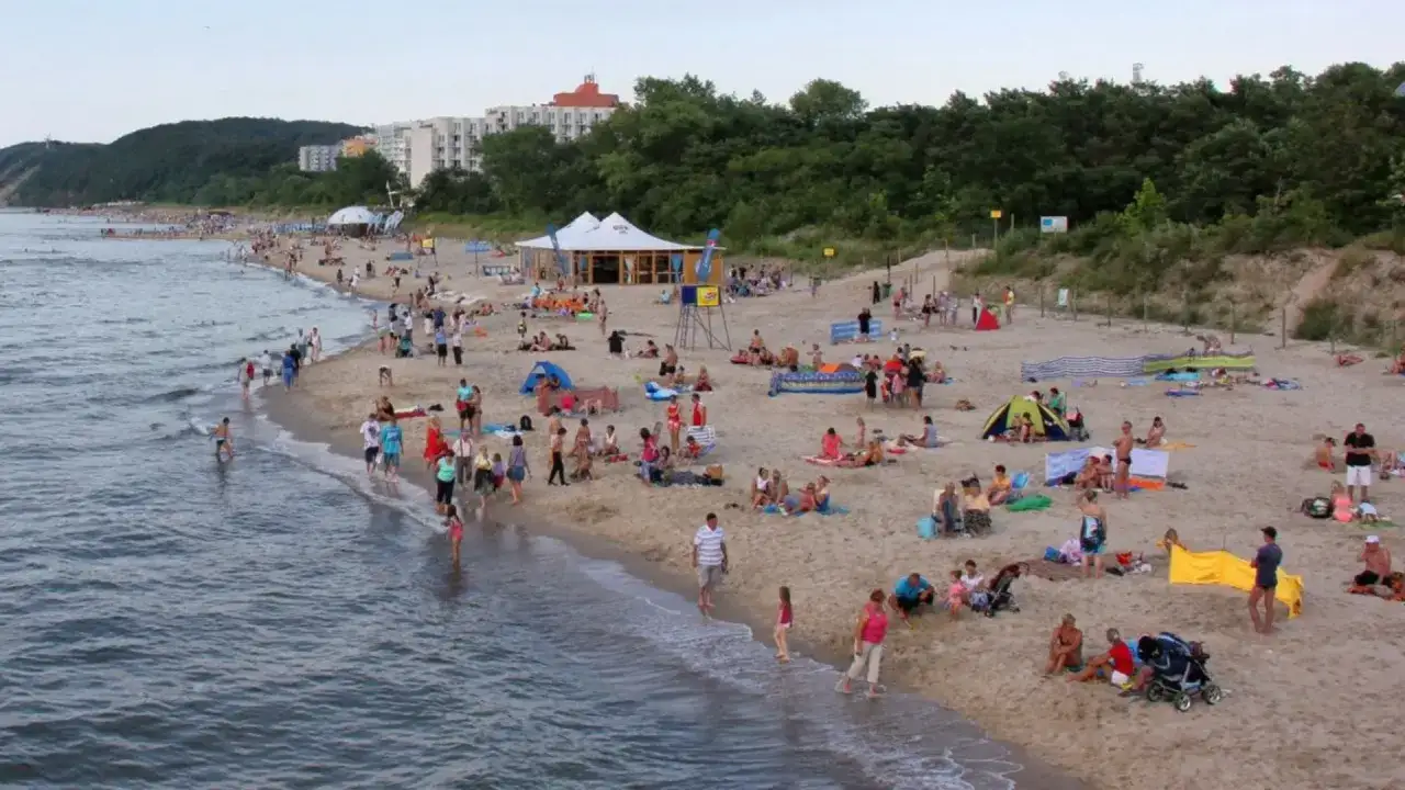 Tętniąca życiem miejscowość nad morzem. Ludzie odpoczywają na plaży, kąpią się w morzu, a w tle widać budynki i zalesione wzg&oacute;rza.