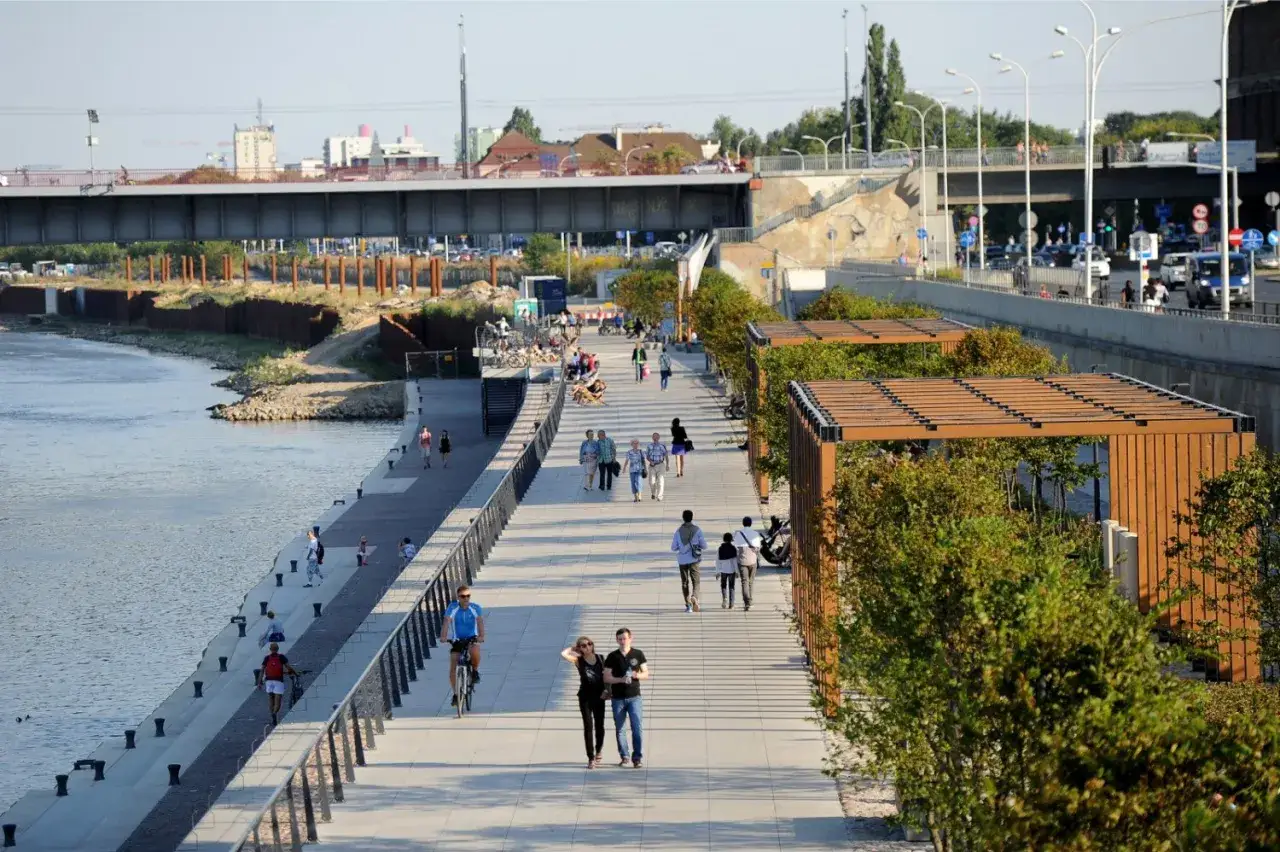 Spacerem nad Wisłą, wśr&oacute;d zieleni i nowoczesnej architektury, odkrywasz fajne miejsca w Warszawie. Ludzie spacerują, jeżdżą na rowerach, ciesząc się dniem.