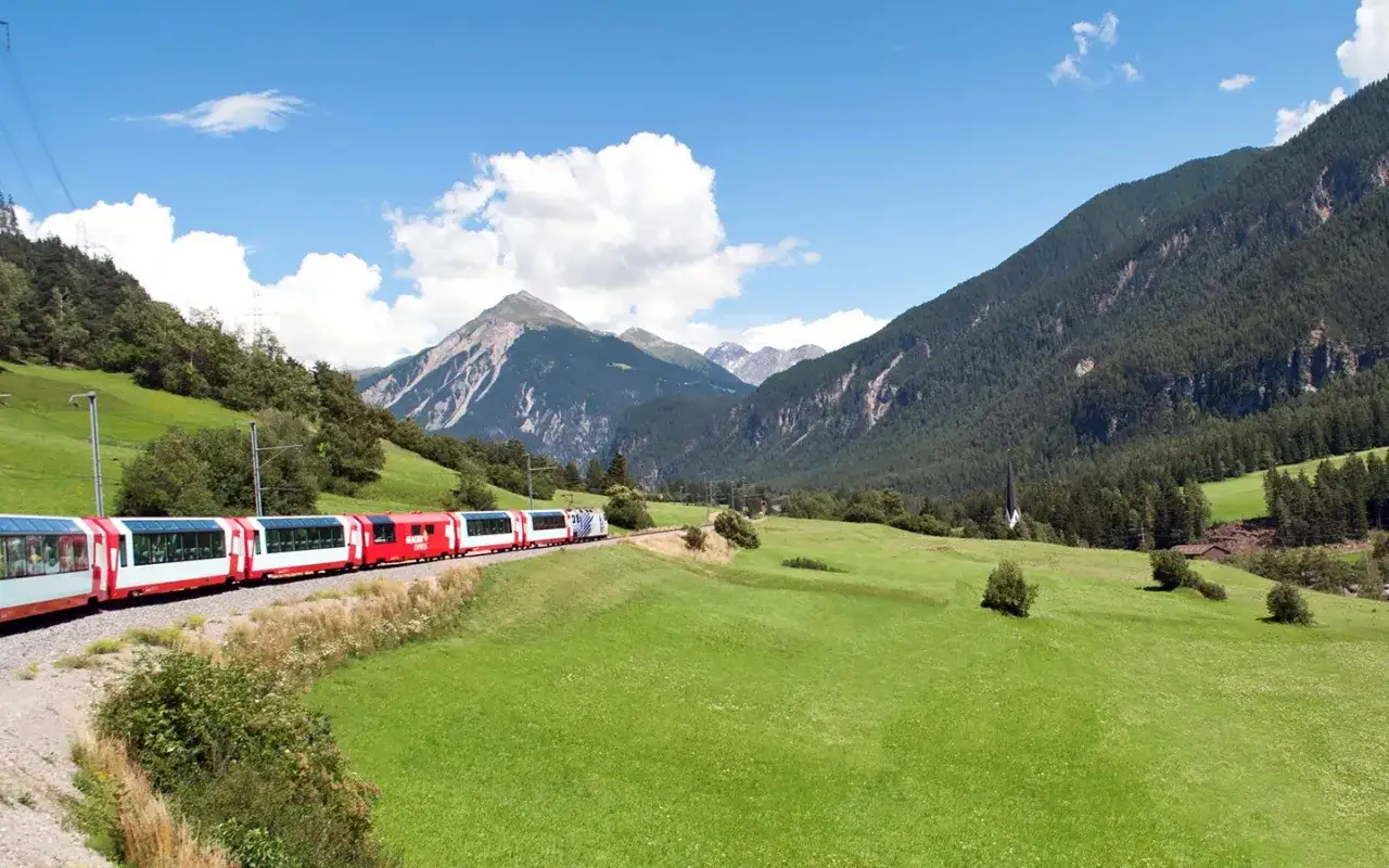 T&aring;g i alplandskap, perfekt f&ouml;r dina interrail tips. Gr&ouml;nskande &auml;ngar, berg och en kyrka.