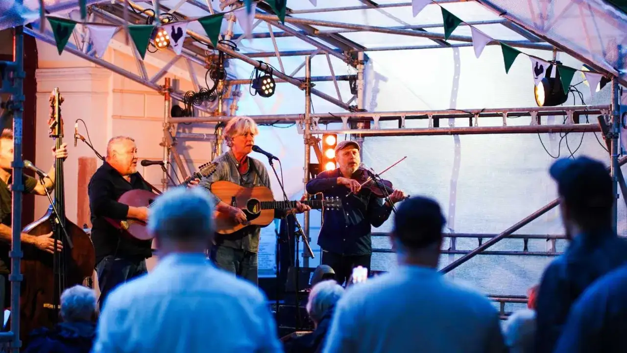En grupp musiker spelar på en utomhusscen. En man spelar gitarr och sjunger, en annan spelar fiol. Publiken njuter av den gratis konsert.