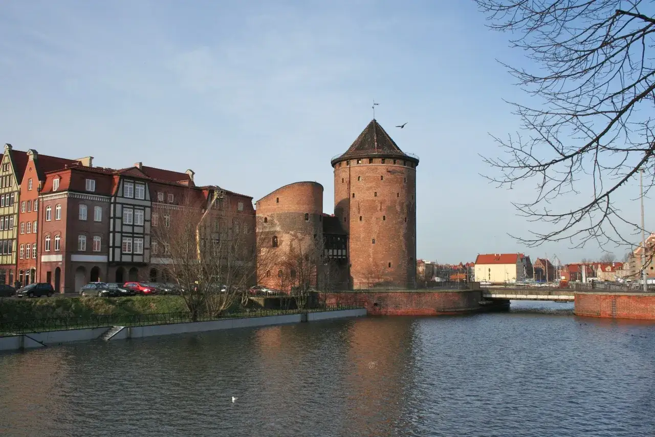 Potężna baszta tajemnic g&oacute;ruje nad wodą, otoczona zabytkowymi kamienicami i mostem.