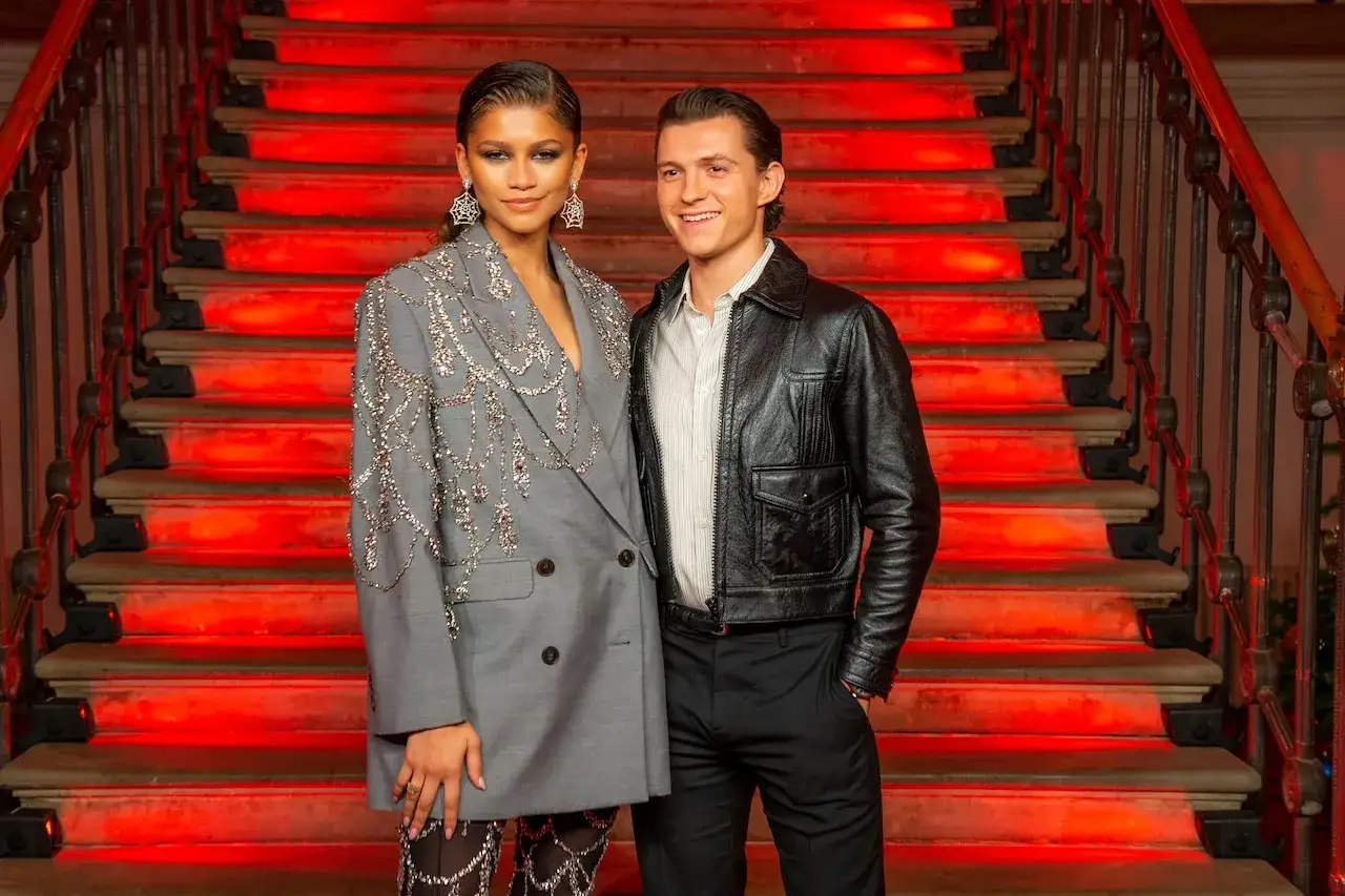 Zendaya i Tom Holland na tle czerwonych schodów. Zendaya w błyszczącej marynarce, Tom w skórzanej kurtce.