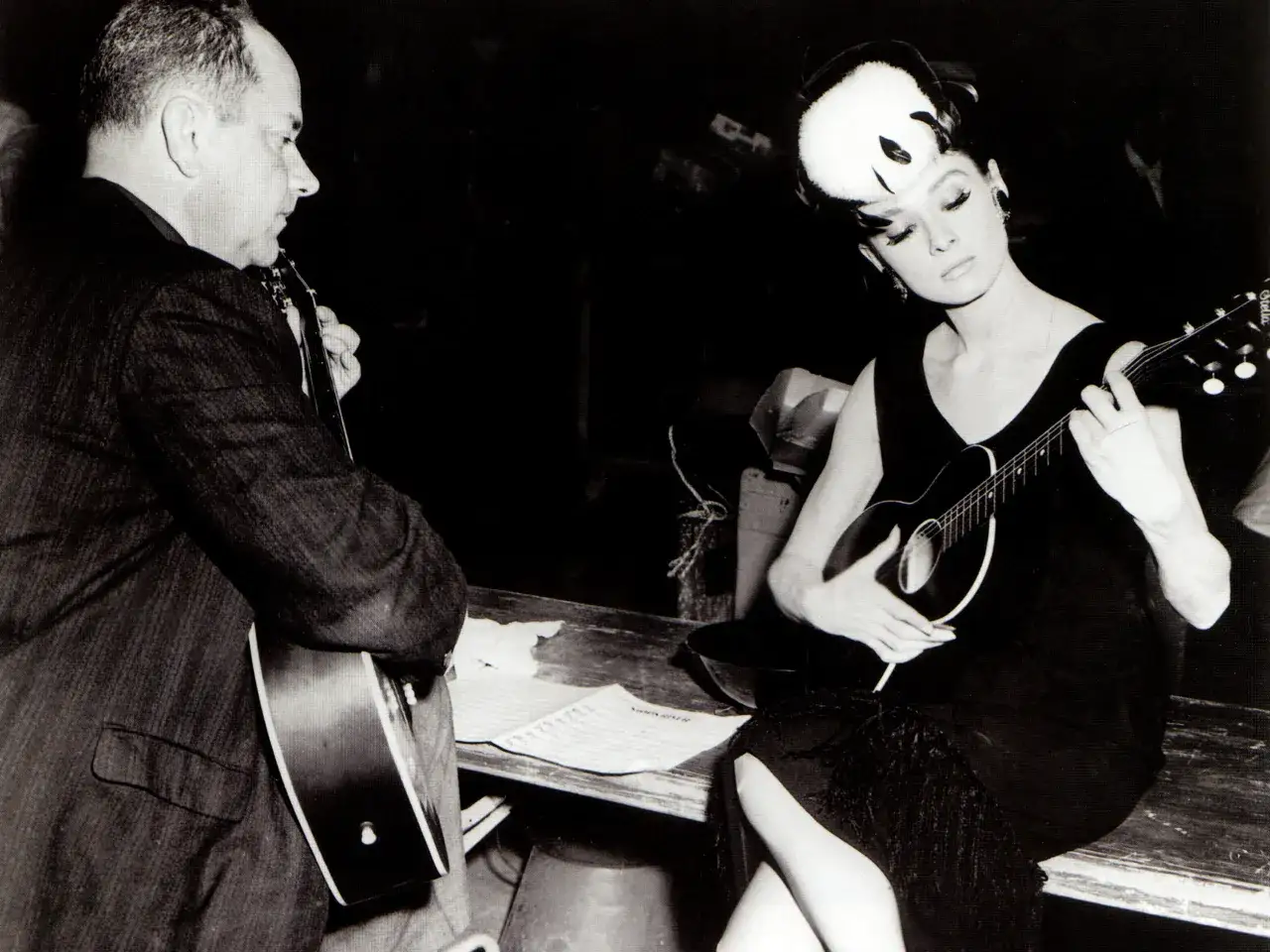 Audrey Hepburn gra na ukulele, a mężczyzna obok na gitarze. W tle nuty z piosenką 
