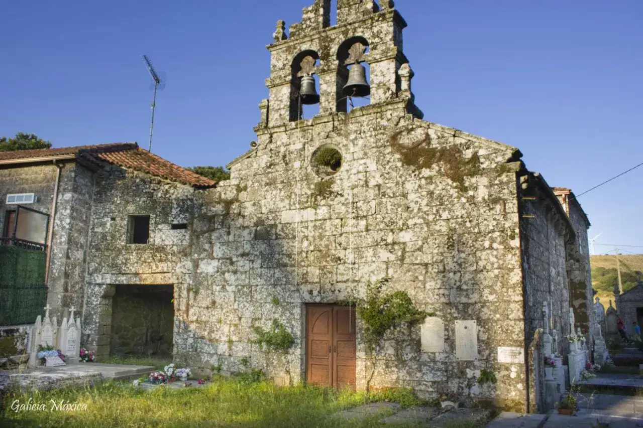 Iglesia de piedra con campanario y campanas, un lugar que ver en Rodeiro. Flores y l&aacute;pidas rodean la entrada.