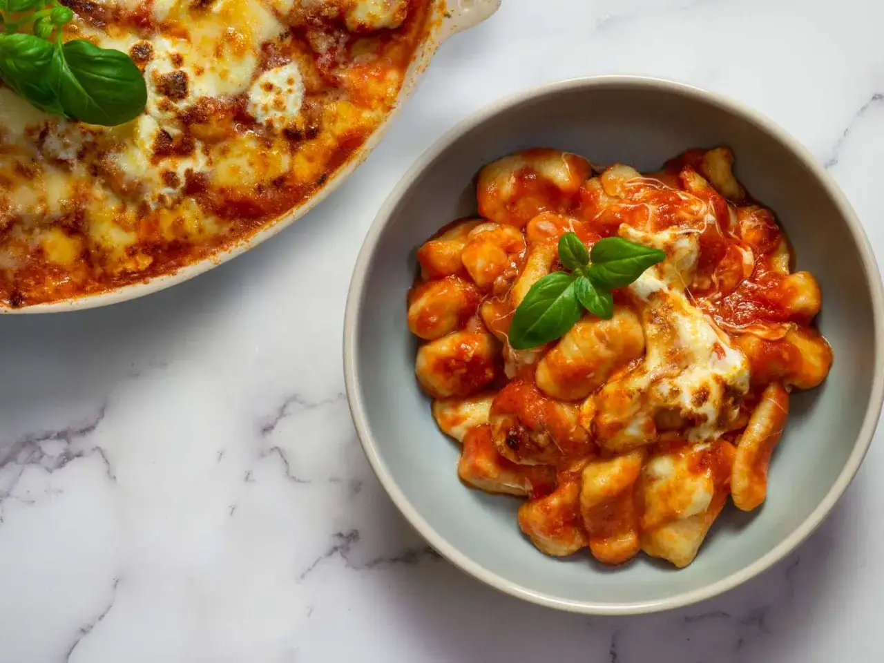 En skål med gnocchi alla sorrentina, täckt av tomatsås och smält ost, med en basilika-kvist. En ugnsform med samma rätt syns i bakgrunden.