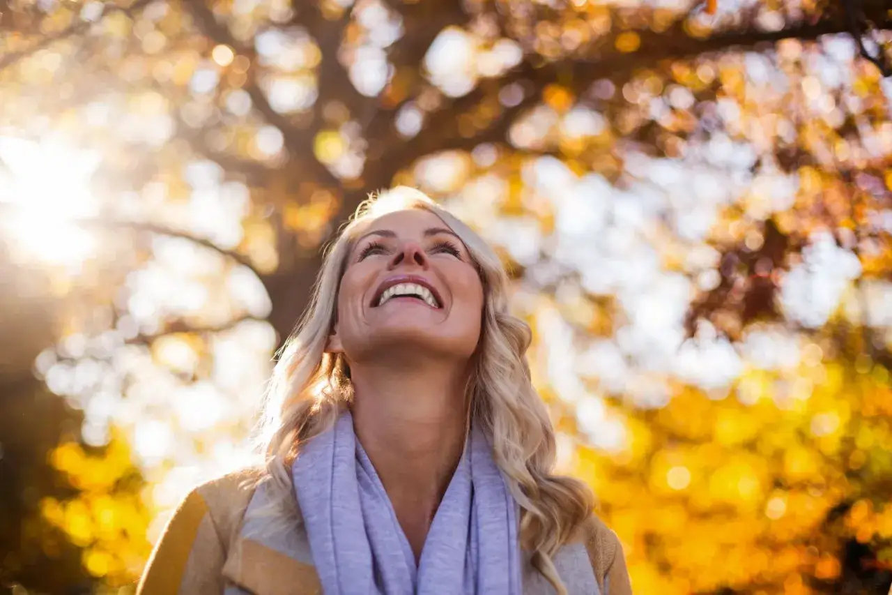 Donna sorride guardando il cielo autunnale, con foglie dorate e luce solare. Forse cerca sollievo dai sintomi cambio stagione.