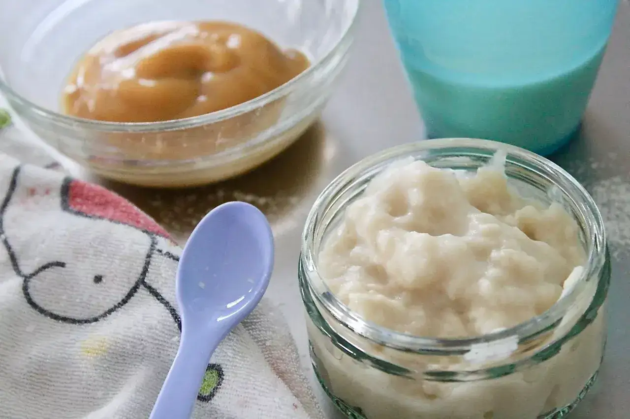 Zwei Schalen mit Babynahrung, eine hell und cremig, die andere braun. Ein L&ouml;ffel und ein Becher mit Milch stehen daneben. Perfekt f&uuml;r den abendbrei mit muttermilch.