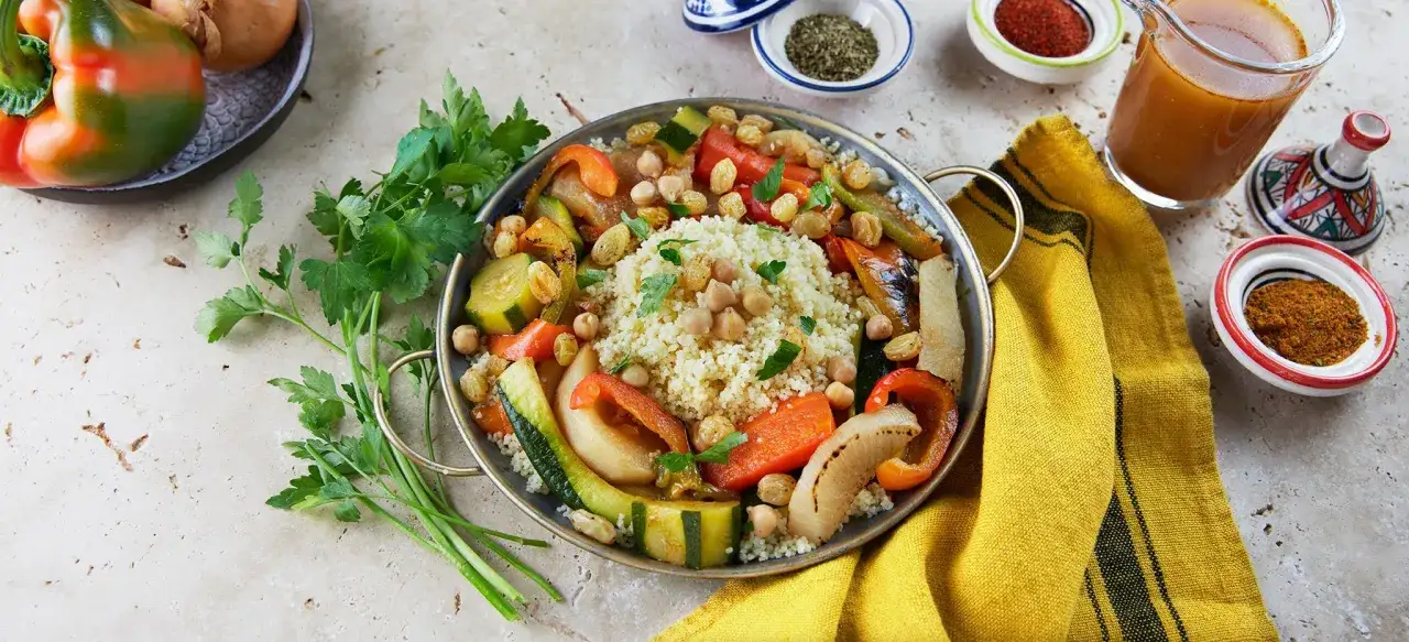 Cuscus con verduras asadas, garbanzos y pasas, servido en un plato met&aacute;lico con perejil fresco.