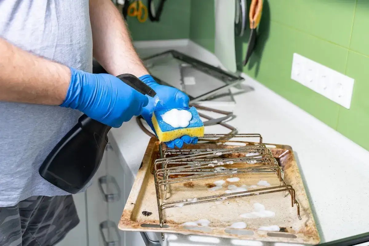 Mit blauen Handschuhen wird ein eingebranntes Backblech gereinigt. Schaum und Sprühflasche sind im Einsatz.